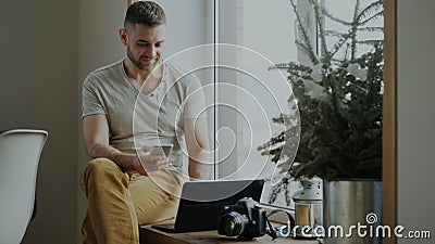 Attractive young man browsing smartphone phone sitting on windowsill with laptop and camera in cafe Stock Photo