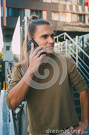 Attractive young man hipster talking on a smartphone while sitting on the steps of a glass business center. Urban fast life Stock Photo