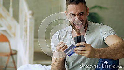 Attractive young man with smartphone and credit card shopping on the internet sit on bed at home Stock Photo