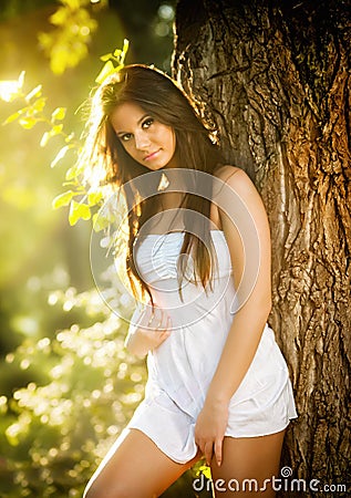 Attractive young woman in white short dress posing near a tree in a sunny summer day. Beautiful girl enjoying the nature Stock Photo