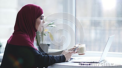 Attraktives moslemisches Mädchen mit hijab auf ihrem Kopf trinkt Cappuccino beim Suchen nach etwas auf ihrem Smartphone stock video Attraktives moslemisches Mädchen mit hijab auf ihrem Kopf trinkt Cappuccino beim Suchen nach etwas auf ihrem Smartphone stock video