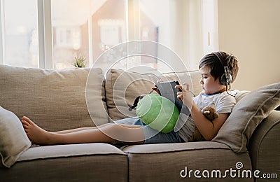 Authentic portrait Kid sitting on sofa watching cartoons on tablet,Yong boy playing game on touch pad, Child lying on couch Stock Photo