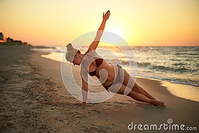 Authentic woman in swimsuit doing yoga vasisthasana on the beach in the morning. Real unretouched shape girl silhouette Stock Photo