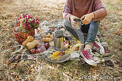 Autumn picnic in the park. The girl holds a cup of tea in her hands. Basket with flowers on a blanket in yellow autumn leaves. Stock Photo