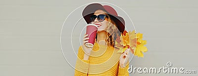 Autumn portrait happy smiling young woman with yellow maple leaves and cup of coffee wearing a sweater and hat on gray Stock Photo