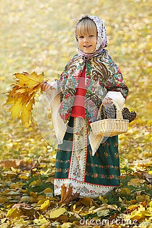 Autumn portrait of the little girl in the traditional russian sarafan and headscarf gathering yellow leaves and pinecones Stock Photo