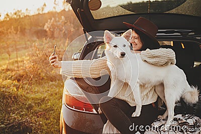 Autumn road trip with pet and travel. Stylish hipster woman taking selfie photo with cute dog in car trunk in sunset light in Stock Photo