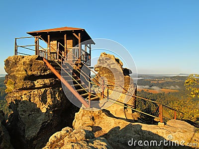 Autumn sunny morning scene. Wooden cabin on main peak of rock as view point, dark sky, autumn mist in valley. Stock Photo