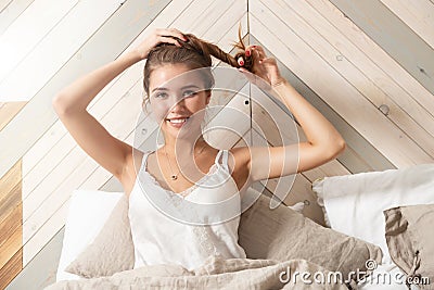 Awakened young smiling woman wearing nightclothes doing hair sitting on bed Stock Photo