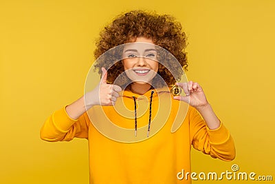 Awesome bitcoin! Portrait of happy satisfied curly-haired woman in urban style hoodie showing thumbs up Stock Photo