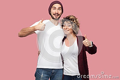 Awesome. Portrait of positive couple of friends in casual style standing, hugging and showing thumbs up with toothy smile, looking Stock Photo