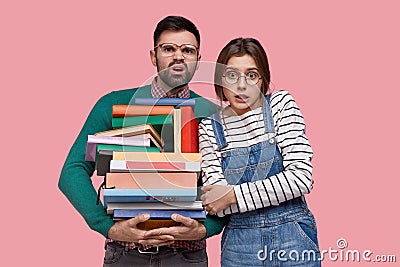 Awkward unshaven young man frowns face, wears big square spectacles, carries many textbooks, young European woman keeps Stock Photo