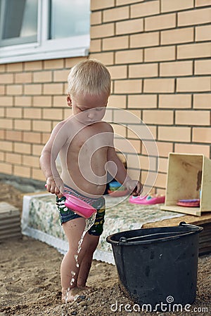 Baby boy with blonde hair plays in the yard of the house in the sand with water. Stock Photo