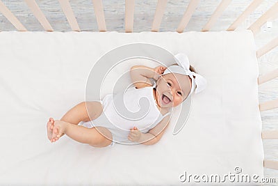 Baby girl 6 months old lies in a crib in the nursery with white clothes on her back and laughs, looks at the camera, baby`s Stock Photo