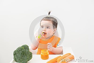 Baby girl in orange bib sitting in a Childs chair eating vegetable puree on white background Stock Photo