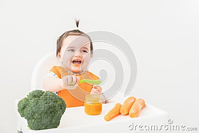 Baby girl in orange bib sitting in a Childs chair eating vegetable puree on white background Stock Photo