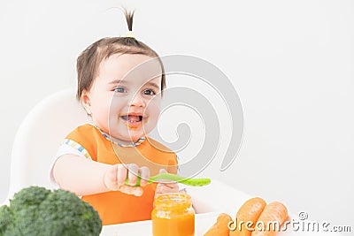 Baby girl in orange bib sitting in a Childs chair eating vegetable puree on white background Stock Photo