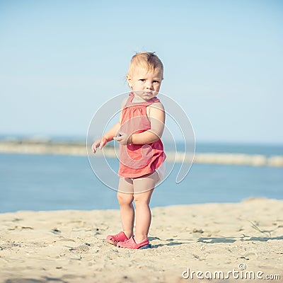 Baby girl in red dress playing on sandy beach near the sea. Stock Photo
