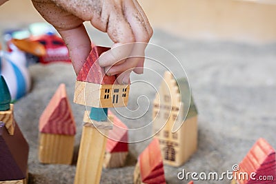 Baby hand with small wooden toy houses on sand. Children game. Five cottages with red roof on grey soil. Modelling Stock Photo