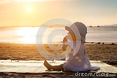 The baby is teething. Everything gnaws, chewing. Infant girl sitting on the tropical sand beach and looking for sunset Stock Photo