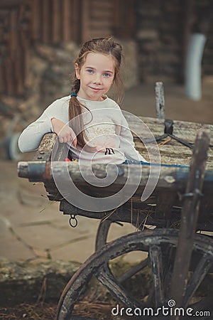 Baby young girl with blue eyes with brunnette plait hair wearing white dress shirt and posing on wooden old style retro wagon cart Stock Photo