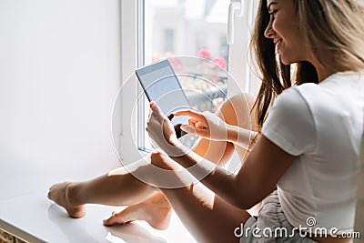 From back pretty girl in shorts on window. She typing on tab and smiling. Stock Photo