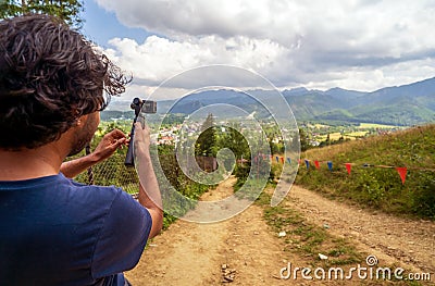 Back or rear view of a male hiker in casual clothes stands in a mountain trail with a sport camera on a stabilizer gimbal and Stock Photo
