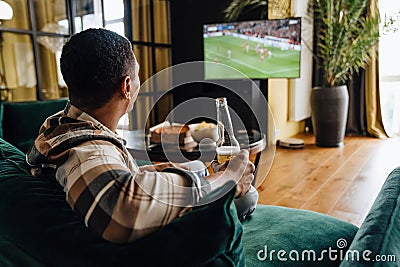 Back view of african man watching football match and drinking beer in front of the TV screen Stock Photo