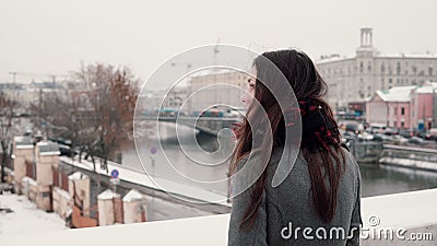 Back view. Attractive young brunette girl standing on the bridge and looks at the snow-covered winter town. Stock Photo