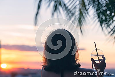 Back view of a beautiful girl with mineral water in a glass against the background of the sea in branches of palm trees. Sunset Stock Photo