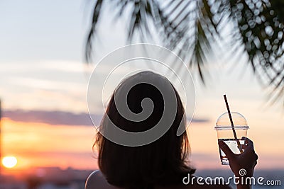 Back view of a beautiful girl with mineral water in a glass against the background of the sea in branches of palm trees. Sunset Stock Photo