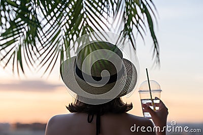 Back view of a beautiful girl with mineral water in a glass in a straw hat against the background of the sea in branches of palm Stock Photo