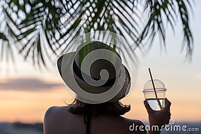 Back view of a beautiful girl with mineral water in a glass in a straw hat against the background of the sea in branches of palm Stock Photo