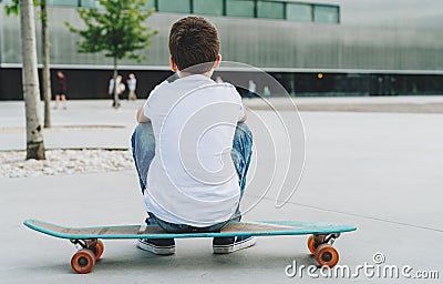 Back view. Boy in white T-shirt and blue jeans, sits on city street on skateboard. Space for text, logo, image. Mock up. Stock Photo