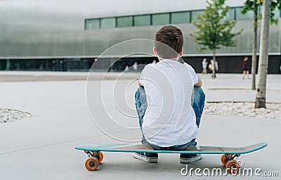 Back view. Boy in white T-shirt and blue jeans, sits on city street on skateboard. Space for text, logo, image. Mock up. Stock Photo