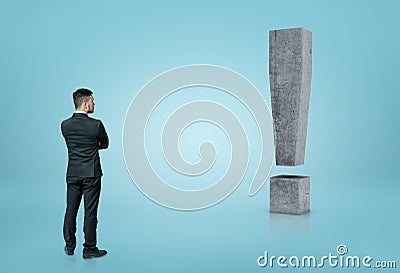 Back view of a businessman looking at big 3D concrete exclamation mark isolated on blue background Stock Photo