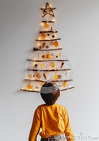 Back view of child looking at handmade craft Christmas tree made from sticks and natural materials hanging on wall. Stock Photo