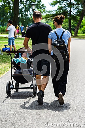 Back view of couple parents holding hands with baby in stroller walking, relaxing and having fun on the alleys of park and gardens Editorial Stock Photo