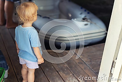 Back view of cute little caucasian toddler boy stand alone in ship garage wooden floor dock and looking on inflatable Stock Photo