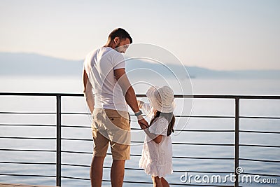 Back view of daughter with her father stand on heigh balcony in front of sea sunset. Dad with his little girl looking on Stock Photo