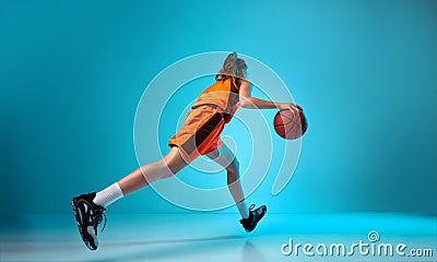 Back view dynamic image of young girl, basketball player dribbling ball against blue studio background in neon light Stock Photo