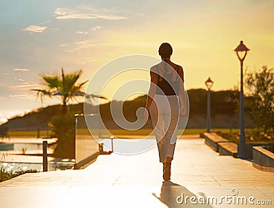 Back view of female silhouette going on evening sunset background of resort hotel. Woman walking by tropic street way with palms Stock Photo