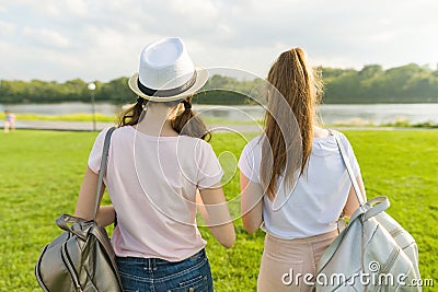 Back view, girl friends are walking in the park in nature. Girls walk along the green lawn, talk, have fun. Stock Photo