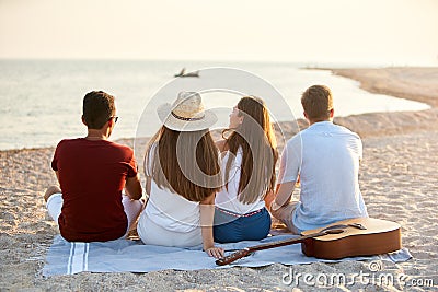 Back view of group of friends sitting together on towel on white sand beach during their vacation and enjoying a sunset Stock Photo