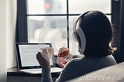 Back view of lonely woman enjoying having breakfast with cup of coffee and croissant, listenning music in headphones working Stock Photo