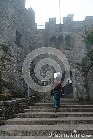 Back view Man with backpack walking alone goes up the stairs. Fortification, old castle. Journey. Mystical atmosphere, fog, white Stock Photo