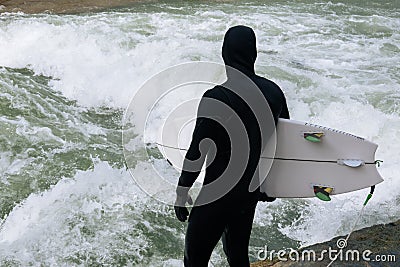 Back view of Man in black wetsuit with white surfboard stains on bank of Eisbach River Germany, Munich, Englischer Stock Photo