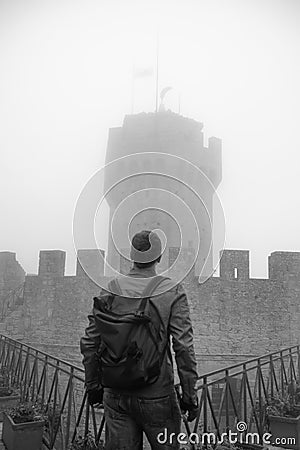 Back view of man wearing leather jacket and backpack. Fortification, old castle on background. Journey. Mystical atmosphere, fog, Stock Photo