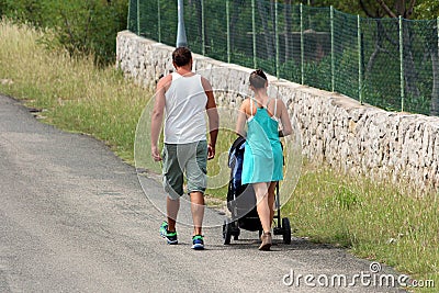 Back view of modern young couple pushing stroller uphill on side of paved road next to uncut grass and traditional stone wall Editorial Stock Photo