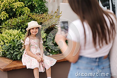 Back view of mom takes photos of her daughter on her phone. Adorable little girl sitting on bench. Focus on kid Stock Photo